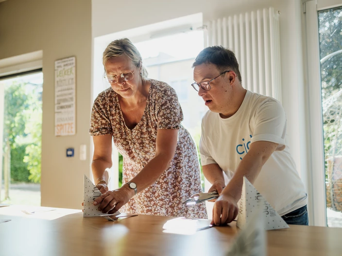 Lehrsituation: Eine Lehrkraft unterstützt einen jungen Mann beim Tischdecken